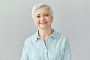 Staršia žena s krátkymi šedivými vlasmi vo svetlom modrom pruhovanom tričku stojí pred jednofarebným svetlým pozadím.