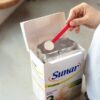 Ruka naberá práškovú Sunar Sensitive 1 dojčenskú výživu 500g červenou lyžičkou z otvorenej krabice.