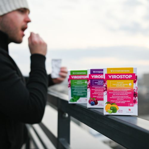 Osoba stojí na balkóne a drží blister s pastilkami, jednu si vkladá do úst. Na zábradlí sú položené tri krabičky pastiliek Virostop.