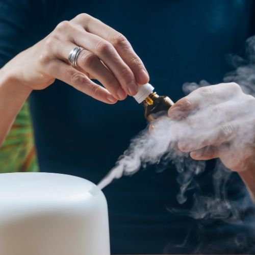 Osoba nalieva esenciálny olej z malej fľaše do difuzéra alebo zvlhčovača vzduchu, z ktorého vychádza viditeľná para.