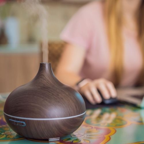 Osoba s počítačovou myšou v pozadí, pred ňou na farebnom stole tmavý difúzér dreveného vzhľadu vypúšťa paru. Spôsob, ako používať esenciálne oleje.