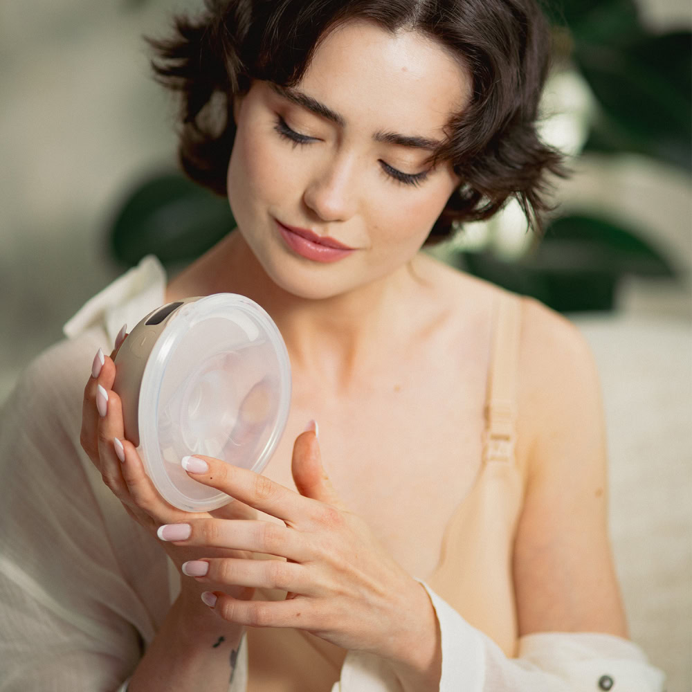 Canpol Babies elektrická odsávačka materského mlieka do podprsenky SmartSense3 Žena v béžovom tope a otvorenej košeli si prezerá Canpol Babies elektrickú odsávačku mlieka, v pozadí je rozmazaná zeleň.