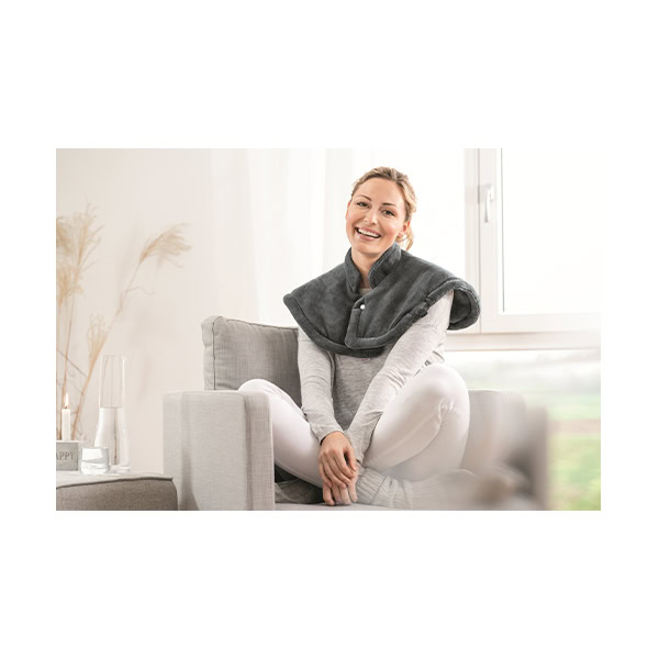 Beurer vyhrievacia podložka na šiju a plecia HK 66_2 Žena sedí so skríženými nohami na sivom kresle v interiéri, má na sebe vyhrievaciu podložku Beurer HK 66 na šiju a plecia.